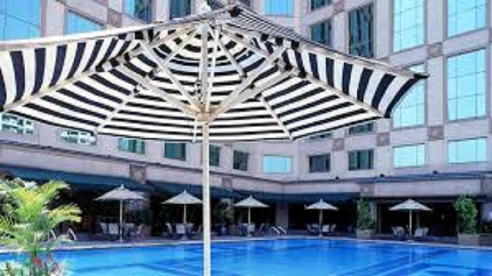 Jw Marriott Kuala Lumpur Hotel Exterior photo The photo shows a swimming pool surrounded by a deck and several striped umbrellas providing shade. The pool has a clear blue water surface, and there are seating areas with lounges or chairs visible near the pool. The surrounding buildings have larg