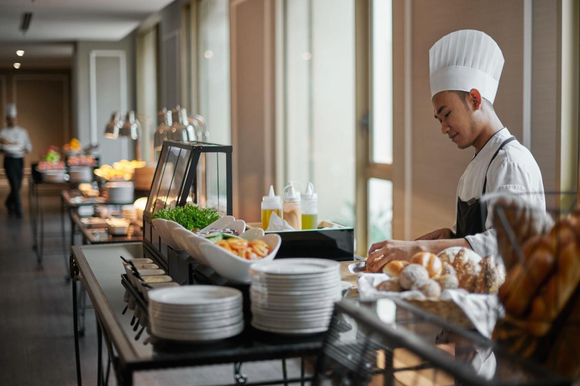 Jw Marriott Kuala Lumpur Hotel Exterior photo The photo depicts a chef working in a buffet setting. He is wearing a chef's hat and uniform, focusing on preparing food. The buffet table is lined with a variety of dishes and ingredients, including salads and bread. There are several plates stacked