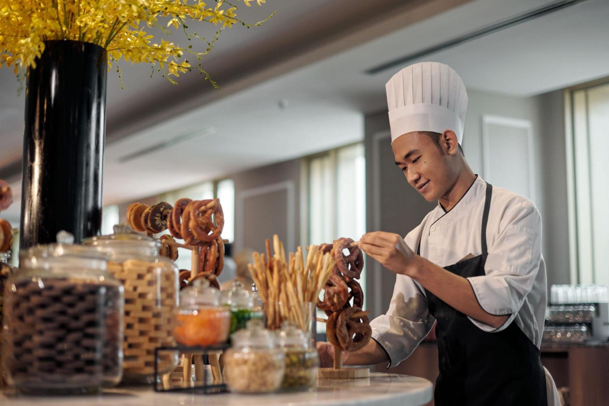 Jw Marriott Kuala Lumpur Hotel Exterior photo The photo shows a young chef in a professional kitchen setting. He is wearing a white chef's hat and a dark apron over a white chef's jacket. The chef is arranging pretzels on a table or display. In the background, there are several jars filled with 