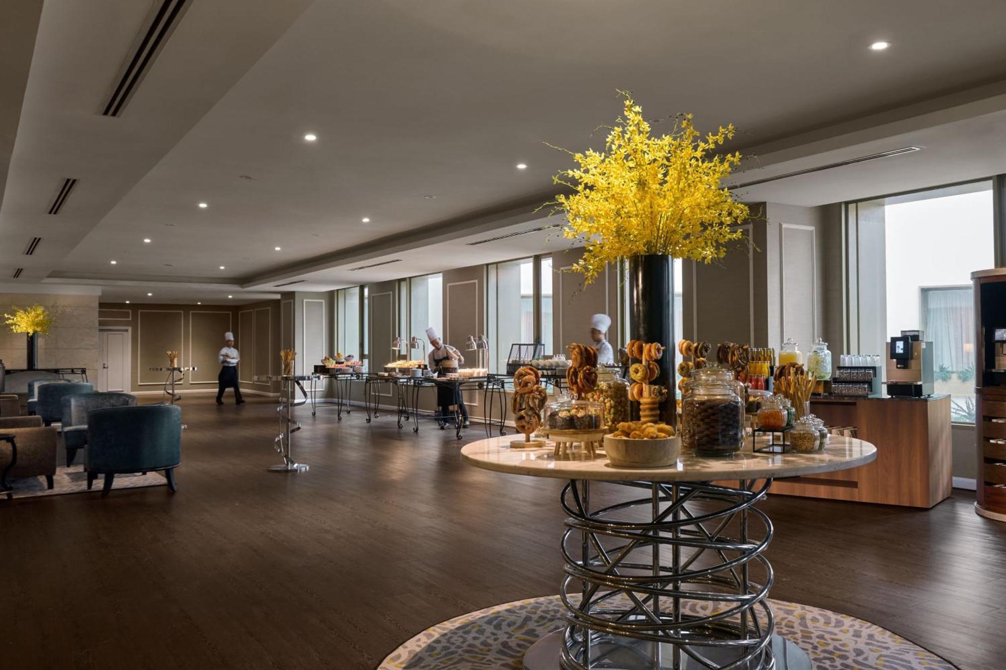 Jw Marriott Kuala Lumpur Hotel Exterior photo The photo shows a modern, elegant dining area, likely set up for a buffet or catering event. In the center, there is a large, round table with a decorative arrangement of yellow flowers, accompanied by various jars and displays of food items. Several
