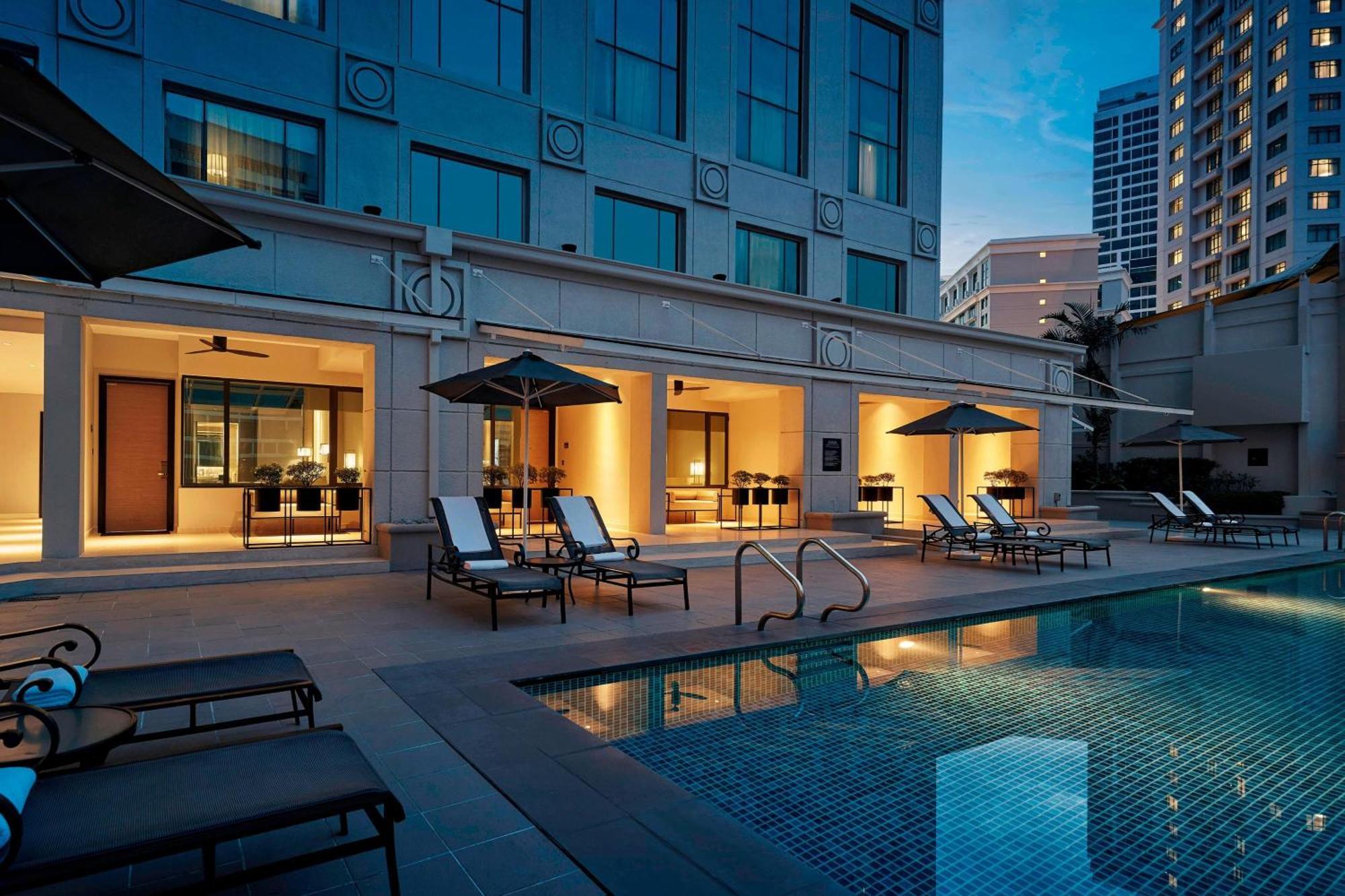 Jw Marriott Kuala Lumpur Hotel Exterior photo The photo depicts a luxurious hotel pool area. The swimming pool features clear water and is surrounded by several lounge chairs under sun umbrellas. In the background, there are large glass windows of the hotel, showing illuminated spaces inside, li