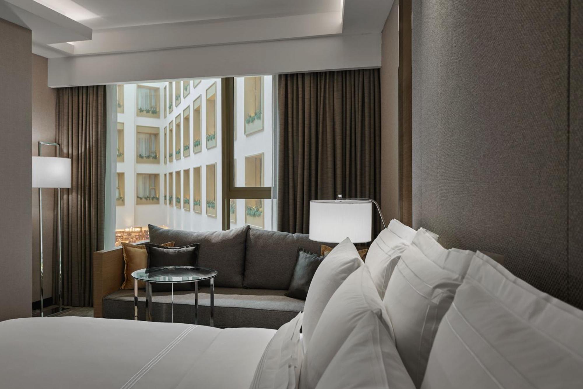 Jw Marriott Kuala Lumpur Hotel Exterior photo The photo features a modern hotel room. In the foreground, there is a large bed with crisp, white linens and multiple pillows. To the side, there is a cozy seating area with a gray sofa and a round glass table. A lamp is situated next to the sofa, pr