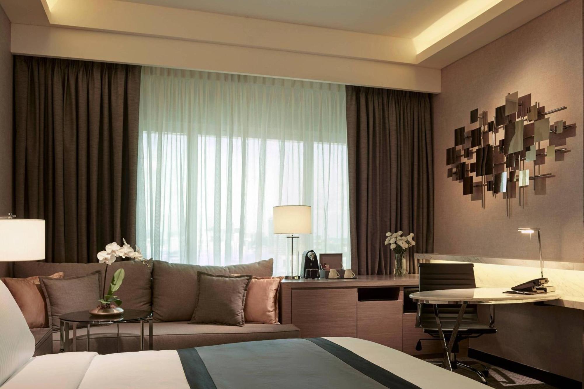 Jw Marriott Kuala Lumpur Hotel Exterior photo The photo shows a modern hotel room interior. There's a comfortable-looking bed with a dark-colored bedspread. A sitting area features a sofa adorned with cushions next to a window, which is partially covered by light, sheer curtains. A lamp is place
