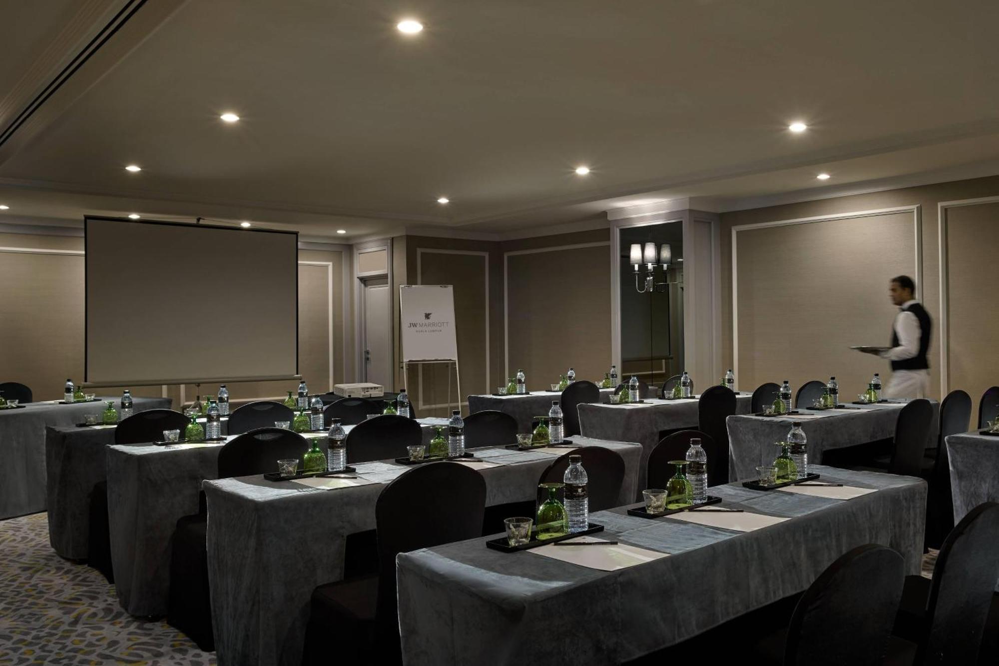 Jw Marriott Kuala Lumpur Hotel Exterior photo The image shows a conference room setup. There are several tables arranged in a U-shape, each covered with a gray tablecloth. On each table, there are bottles of water and other supplies placed strategically. The room is softly lit with ceiling light