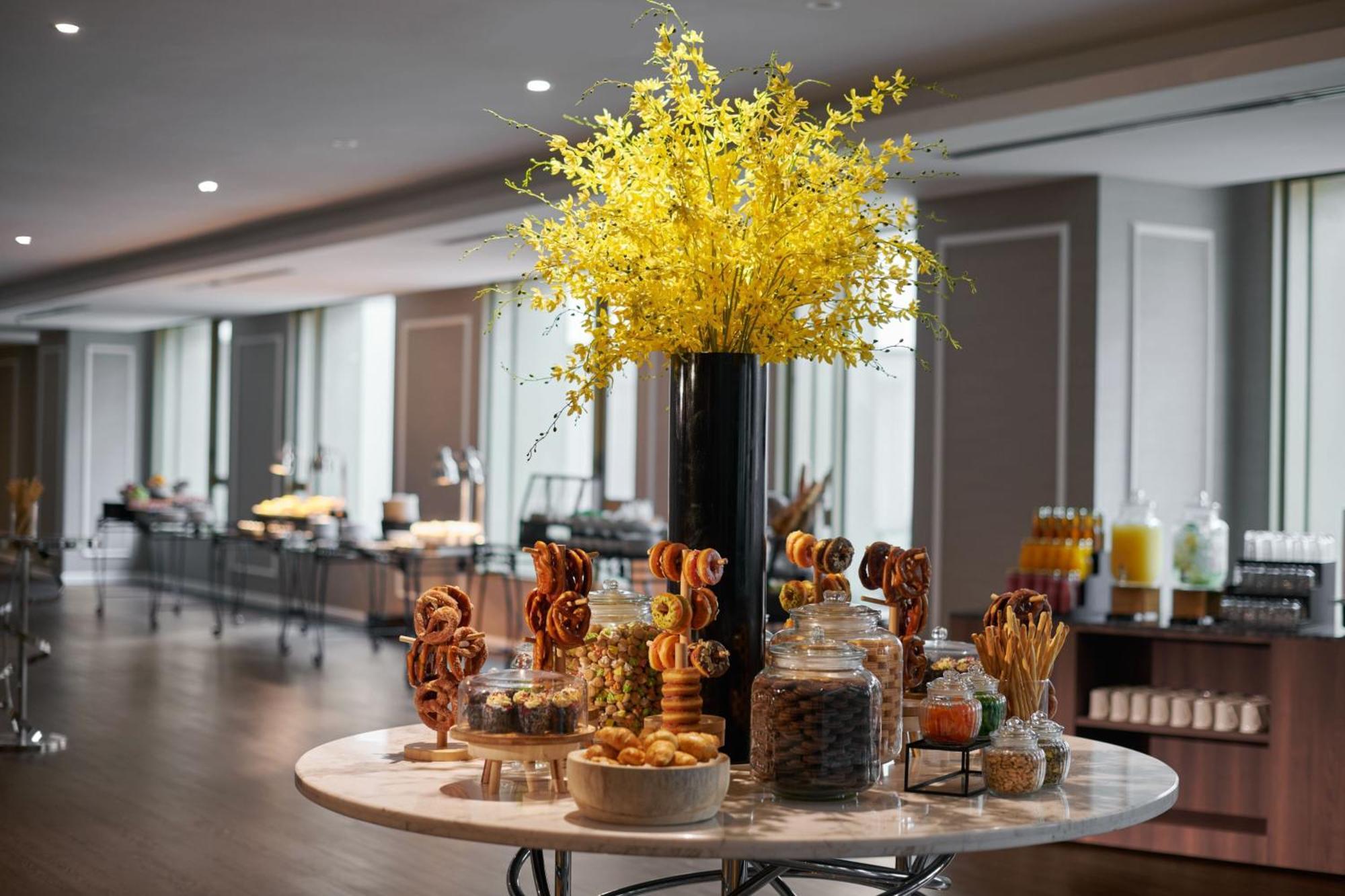 Jw Marriott Kuala Lumpur Hotel Exterior photo The photo depicts a beautifully arranged display in a dining or banquet setting. At the center is a round table featuring an elegant floral arrangement with bright yellow flowers in a tall vase. Surrounding the flowers are various jars filled with sw