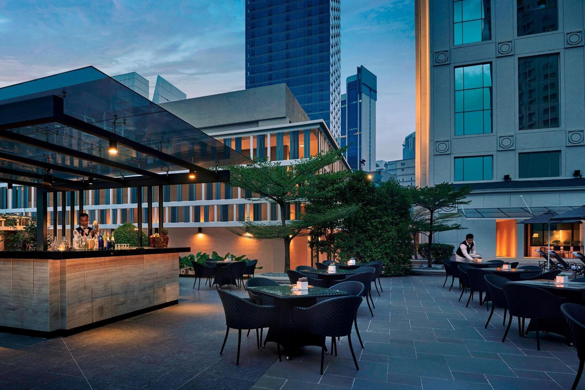 Jw Marriott Kuala Lumpur Hotel Exterior photo The photo depicts a modern outdoor terrace or rooftop bar. It features a stylish bar area with an elegant glass structure, set against a backdrop of tall buildings. There are several tables with dark, woven chairs, arranged for guests to sit. The atm
